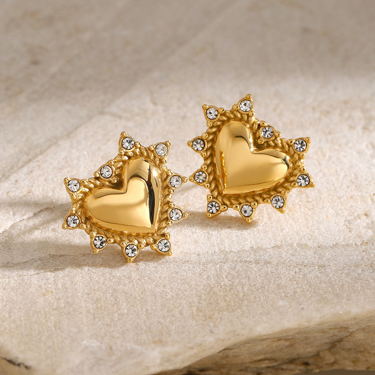 Gold heart-shaped stud earrings with sparkling crystal accents on a neutral stone background – luxury jewelry by Arge Steel